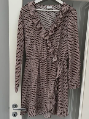 Rosa klänning från Vila, dam stl M - Gammelrosa kort klänning från Vila, dam stl M, med svart prickigt mönster. Klänningen har volangdetaljer längs v-ringningen och omlott ner till midjan. 