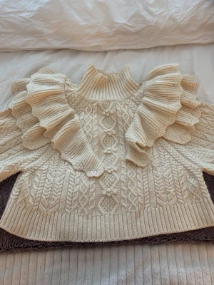 Beige stickad polotröja med volang Zara - Superfin beige stickad polotröja från Zara med breda volangdetaljer över axlarna och snyggt kabelstickat mönster. Tröjan har ribbad polokrage och långärmade ärmar. Perfekt för dig som gillar unika detaljer och vill ha en cozy look.