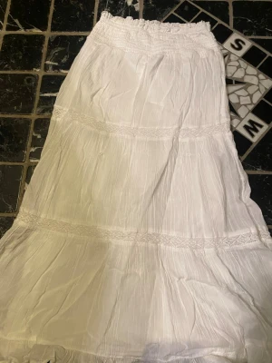 Vit långkjol med volangdetaljer - Säljer en vit långkjol från Gina Tricot med smock i midjan och flera volangdetaljer. Kjolen är gjord i ett tunt, luftigt tyg som känns perfekt för varma dagar. Enkel att matcha och ger en bohemisk vibe.