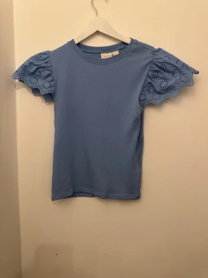 Blå topp med broderade volangärmar - Supersöt blå topp från Gina Tricot med korta volangärmar i broderad bomull. Ärmarna har fina hålmönster och ger en extra touch till en annars enkel och stilren topp. Perfekt till jeans eller kjol för en fräsch look.