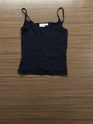 Svart linnetopp från Gina Tricot - En enkel svart linne från Gina Tricot med smala axelband. Toppen är rak i modellen och tillverkad i mjuk bomull, perfekt för varma dagar eller som lager under andra plagg.