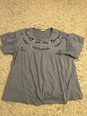 Blå broderad blus från mango - Supersöt ljusblå blus från Mango med broderade blommor i svart och vitt över bröstet. Modellen har korta puffärmar med volangdetalj och rund halsringning. Använd några gånger. 