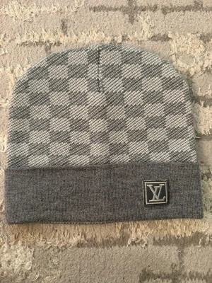 Gråvit ruttig Luice vitton vest - Säljer en stilren väst från Louis Vuitton i grått och vitt med rutigt mönster. Västen har en bred grå kant nedtill och ett broderat LV-emblem framtill. Materialet är stickat och känns mjukt mot huden. Perfekt för dig som gillar exklusiva märken och klassisk design.