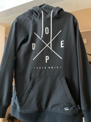Skidjacka Svart hoodie från Dope - Svart hoodie från Dope med vit grafisk text och korsade linjer på bröstet. Stor känguruficka framtill, vita dragsnören i huvan, skaljacka perfekt för backen och afterski. 