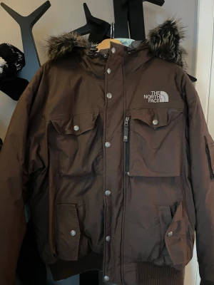 Brun vinterjacka från The North Face - Säljer en brun dunjacka från The North Face med stor huva och fejkpäls. Jackan har flera fickor med knappar och dragkedja, samt ribbade muddar. Perfekt för kalla dagar och riktigt snygg streetstil.