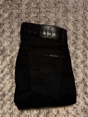 Nudie Jeans - Säljer ett par svarta skinny jeans från Nudie Jeans med klassisk femficksdesign och snygga svarta sömmar. Jeansen har en stilren look med svart läderpatch i midjan och är tillverkade i stretchig bomull för extra komfort.