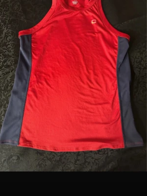 Röd träningslinne från CLN Athletics  st 38  - Snygg röd träningslinne från CLN Athletics i storlek M. Linnet har marinblå paneler på sidorna och CLN-logga både fram och bak. Tillverkat i lätt och ventilerande polyester, perfekt för gym eller löpning. Sportig design med brottarrygg och tryckta detaljer.