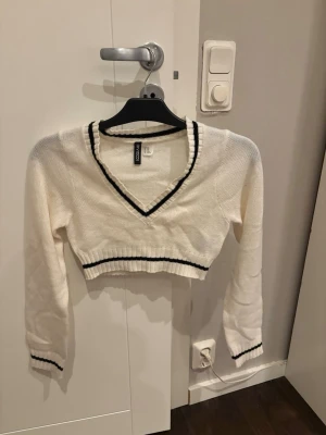 Vit croppad stickad tröja H&M - Säljer en vit croppad stickad tröja från H&M med svarta ränder vid v-ringad hals, midja och ärmslut. Tröjan har lång ärm och ribbade detaljer som ger en sportig och fräsch look. Perfekt att styla med jeans eller kjol.