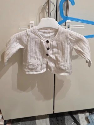 Vit stickad kofta från Kappahl baby - Supersöt vit stickad kofta från Kappahl baby i storlek 80. Koftan har dekorativa bruna knappar framtill, två små fickor och ett diskret hålmönster. Perfekt till kyliga dagar och enkel att matcha med andra plagg.