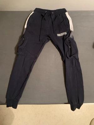 Mörkblå Hoodrich cargopants XS - Säljer ett par mörkblå cargopants från Hoodrich i storlek XS. Byxorna har vita sidopaneler, snörning i midjan och flera fickor med dragkedja. Materialet är mjukt och skönt, troligen bomull eller polyester. Hoodrich-logga tryckt på benet och framtill.