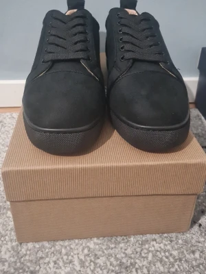 Svarta sneakers från Christian Louboutin - Säljer ett par svarta sneakers från Christian Louboutin i mocka med klassisk röd sula och diskret broderad logga på hälen. Skorna har rund tå, snörning och en stilren look som passar till det mesta. Box och dustbag medföljer.