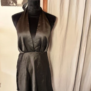 Svart omlottklänning med halterneck - Snygg svart omlottklänning med halterneck och djup v-ringning. Klänningen är kort och har en lätt glansig yta som ger en elegant känsla. Materialet känns som satin eller polyester och den stängs med dragkedja baktill. Perfekt för dig som gillar stilrena och trendiga plagg.