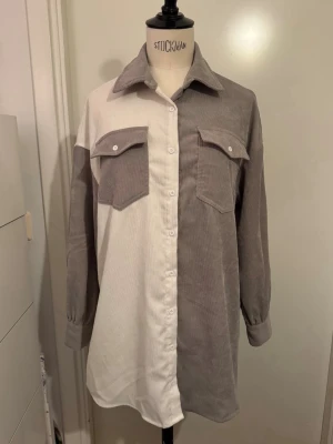 Tvåfärgad overshirt  - Säljer en ny oversized skjorta i mjuk kvalitet med tvåfärgad design i beige och vitt. Skjortan har stora bröstfickor med lock, klassisk krage och knappar hela vägen framtill. Stl 12-13 år