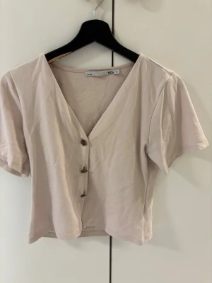Beige kortärmad blus från 157 - Säljer en ljusrosa blus från 157 i storlek S. Toppen har v-ringning, tre dekorativa knappar framtill och är kortärmad med en croppad passform