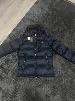 Mörkblå dunjacka från Polo Ralph Lauren - Säljer en mörkblå dunjacka från Polo Ralph Lauren med hög krage och två dragkedjeförsedda fickor. Jackan har det klassiska röda Polo-logot på bröstet och en clean, quiltad design. Perfekt för kalla dagar och riktigt snygg till streetwear-stilen.