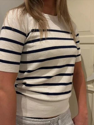 Vit och marinblå randig topp - Säljer en vit topp med marinblå horisontella ränder. Modellen har rund hals, korta ärmar och ribbad kant nedtill och vid ärmslut. Materialet känns mjukt och lätt. Passformen är normal och toppen är enkel att matcha med jeans eller kjol.