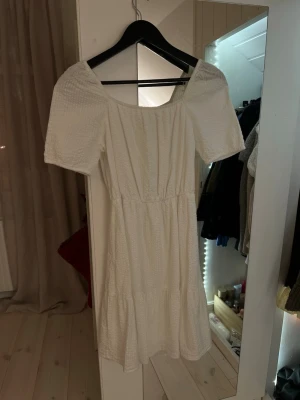 Vit klänning med knyt från Lindex - Säljer en vit klänning från Lindex barn med korta ärmar och bäckelbölja struktur i tyget. Baksidan har en öppen rygg med knyt och rosett. Klänningen har en lätt volang nedtill. Använd 1 gång på en examen. 