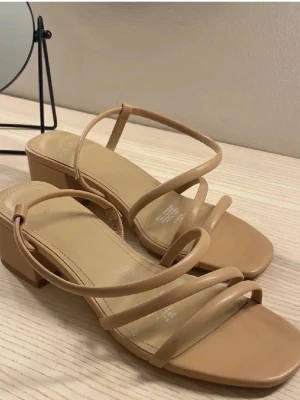 Beige sandaler med blockklack från H&M - Säljer ett par stilrena beige sandaler från H&M med tunna remmar och fyrkantig tå. Skorna har en låg blockklack och öppen tå, perfekta till sommaren. Tillverkade i syntetmaterial med minimalistisk design.