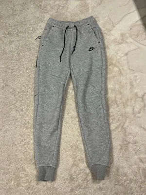 Grå mjukisbyxor från Nike med snörning - Säljer ett par grå mjukisbyxor från Nike i storlek S. Byxorna har svarta snören i midjan, muddar vid bensluten och två fickor fram. Klassisk Nike-logga broderad på vänster sida. Perfekta för chill eller träning.