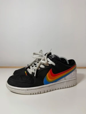 Nike SB Dunk Low Pro Polaroid  - Säljer ett par Nike SB Dunk Low Pro sneakers i svart och vitt med en färgglad regnbågs-swoosh i rött, orange, gult och blått. Skorna har vita snören, perforerad tå och klassisk låg siluett. Ovandelen är i skinn och syntet, perfekt för dig som gillar statement-detaljer.