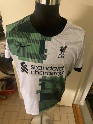 Liverpool FC Salah 11 Nike fotbollströja - Säljer en Liverpool FC fotbollströja med M. Salah och nummer 11 på ryggen. Tröjan är vit med gröna och svarta grafiska mönster, har svarta detaljer vid krage och ärmslut samt Nike-logga på bröstet. Perfekt för dig som älskar fotboll och vill ha en snygg supportertröja.