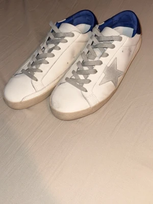 Golden Goose Superstar sneakers vit/blå - Golden Goose Superstar sneakers i vitt skinn med grå mockastjärna på sidan, blå detaljer vid hälen och grå snörning. Platt sula och rund tå. Snygg och klassisk modell med box medföljer. Perfekt för dig som gillar streetstyle och coola detaljer. Aldrig använda bara testat nån gång. Priset går att diskuteras vid snabb affär