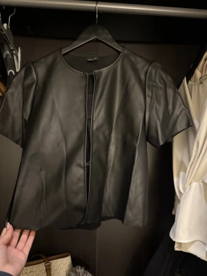 Svart kortärmad skinntröja Gina Tricot - Kortärmad svart tröja i skinnimitation från Gina Tricot. Tröjan har rund halsringning, diskreta knappar framtill och en stilren, minimalistisk look. Perfekt för dig som vill ha en edgy vibe till din outfit.