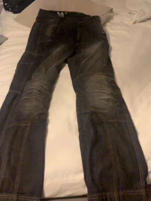 Svarta jeansbyxor med kevlar från Held - Säljer ett par svarta jeansbyxor från Held med coola sömdetaljer och förstärkningar på knäna. Byxorna är tillverkade i 100% bomull med foder i kevlar, vilket gör dem extra slitstarka. Perfekt för dig som vill ha en tuff och funktionell look.