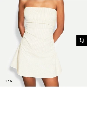 Axelbandslös kortklänning - Supersnygg axelbandslös kortklänning i vit med diskreta smala ränder. Klänningen har en figurnära överdel och utställd kjol som ger en fräsch och somrig look. Perfekt för dig som gillar minimalistisk stil och vill ha något lätt och luftigt.