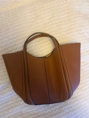 Brun handväska Zara - Snygg brun handväska i skinn med två handtag och avtagbar axelrem. Väskan har en stilren design med dekorativa sömmar och praktiskt innerfack med dragkedja. Kommer med ett långt band som man kan sätta på väskan💖