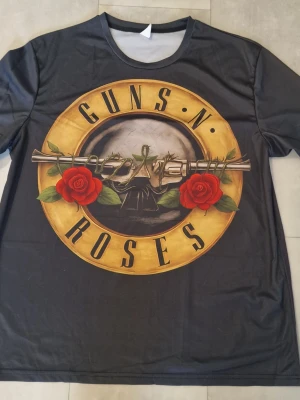 Guns N' Roses svart t-shirt - Svart t-shirt med stort Guns N' Roses-tryck både fram och bak. Trycket har gul cirkel, röda rosor och metalliska revolvrar. Rund hals och korta ärmar. Materialet känns lätt och syntetiskt, perfekt för dig som vill sticka ut med en ikonisk bandtröja.