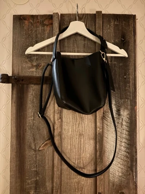 Svart axelväska i skinnimitation - Snygg svart axelväska med minimalistisk design och justerbar axelrem. Väskan har en stilren form och stängs med en enkel karbinhake. Perfekt för dig som gillar en clean och modern look.