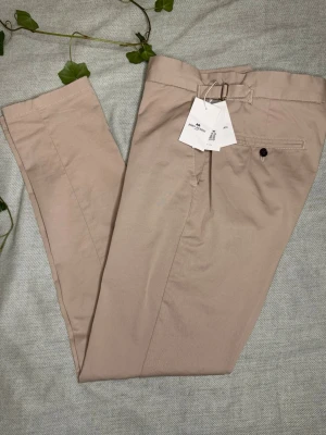 Beige chinos från John Henric - Snygga beige chinos från John Henric med klassisk passform och diskret bältesdetalj i midjan. Byxorna har raka ben, knappstängning bak och är tillverkade i mjuk bomull. Perfekta för en clean och stilren look. 