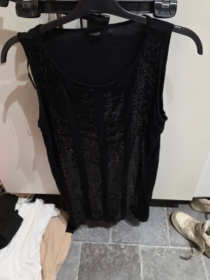 Svart paljettopp från Lindex Generous - Säljer en svart ärmlös topp från Lindex Generous i storlek L. Toppen har breda axelband och är dekorerad med vertikala paljettränder framtill som ger ett glittrigt intryck. Tillverkad i mjuk viskos som känns skön mot huden. Perfekt för dig som vill sticka ut lite extra.