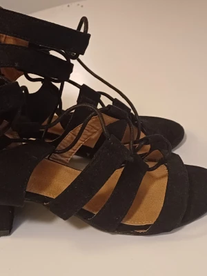 Svarta sandaler med snörning och klack - Säljer ett par svarta sandaler med breda remmar och snörning som går upp runt ankeln. Skorna har en låg klack och öppen tå, vilket ger en snygg och trendig look. Materialet är mocka och sulan är platt framtill. Perfekt för dig som gillar stilrena detaljer.