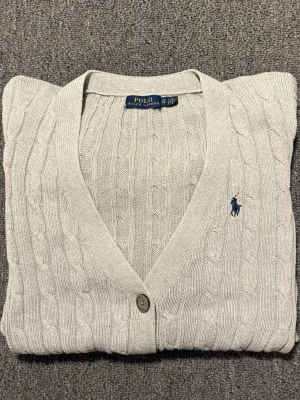 kabelstickad kofta Polo Ralph Lauren - Snygg beige kabelstickad kofta från Polo Ralph Lauren i storlek XS. Koftan har v-ringning, knappar framtill och två små fickor. Klassisk blå broderad logga på bröstet. Perfekt för lager-på-lager och chill stil.