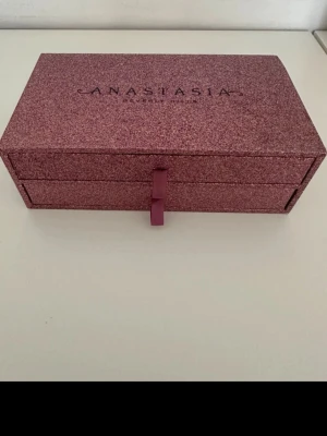 Glittrig sminkbox Anastasia Beverly Hills - Säljer en lyxig sminkbox från Anastasia Beverly Hills i glittrigt rosa/lila material. Boxen har två utdragbara fack och en snygg satinband-detalj framtill. Perfekt för att förvara smink och borstar på ett stilrent sätt.