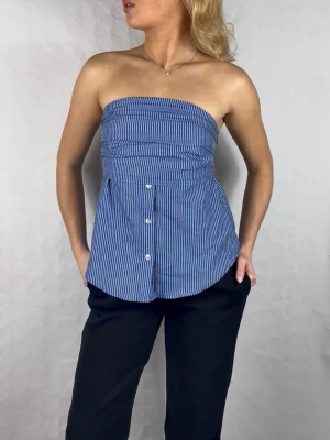 Blårandig tubtopp i skjortmaterial - Säljer denna supersnygga blå- och vitrandiga tubtopp! 💙 Den har en cool 'skjort-look' med knappar framtill och rynkad detalj över bysten vilket ger en jättefin passform. Perfekt att styla till ett par kostymbyxor för en 'officecore'-look eller med baggy jeans för en mer avslappnad stil. 