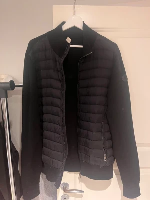 Svart Moncler kofta med dragkedja - Säljer en svart Moncler kofta med quiltad front och stickade ärmar. Tröjan har dragkedja framtill och två sidofickor med dragkedja. Klassiska Moncler-logga på ärmen fast i svart och stilren design som passar perfekt till höst och vinter. Storlek XXL