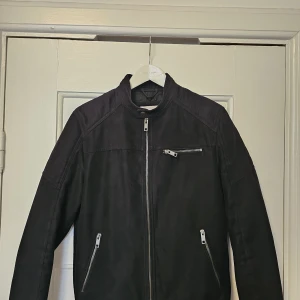 Svart skinnjacka med dragkedjor - Snygg svart skinnjacka med ståkrage och tre silverfärgade dragkedjor framtill. Jackan har en stilren design och långärmade ärmar, perfekt för dig som gillar en cool och klassisk look.