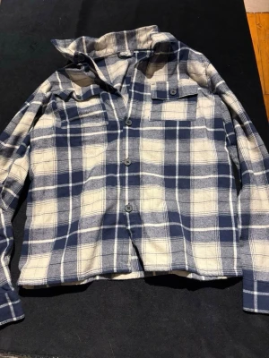 Rutig overshirt från KappAhl blå/vit - Snygg rutig overshirt från KappAhl i blått och vitt. Jackan har klassisk krage, knappstängning framtill och en bröstficka med knapp. Perfekt att slänga på över en hoodie eller t-shirt för en chill look.