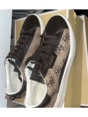 Helt nya bruna sneakers från Guess med  - Snygga bruna sneakers från Guess med klassiskt logomönster på sidan och vita sulor. Skorna har snörning och detaljer i både skinn och textil. Perfekta för dig som vill ha en stilren men ändå trendig look. Helt oanvända och fri frakt!