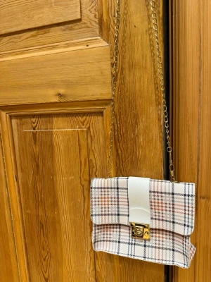 Rutig axelväska med kedja - Säljer en snygg axelväska i rutigt mönster med vita, svarta och röda toner. Väskan har guldfärgad kedja, vit detalj på locket och ett guldfärgat lås framtill. Materialet känns som syntet och väskan har en praktisk fick min bak. Perfekt för dig som vill ha en trendig accessoar. Tror att den är från SHEIN men har använt max 3 ggr. 