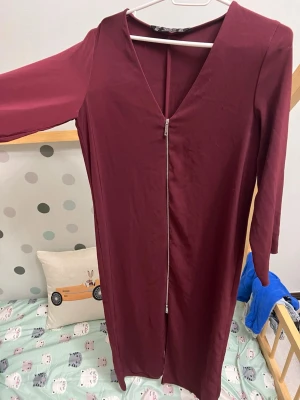 Vinröd långklänning med dragkedja - Säljer en vinröd långklänning från Tailor med v-ringad hals och lång ärm. Klänningen har en snygg dragkedja framtill som detalj och är gjord i ett mjukt polyestermaterial. Perfekt för dig som gillar stilrena och enkla plagg med en twist.