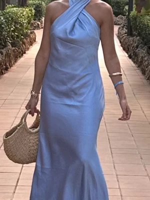 Ljusblå långklänning med omlott - Säljer en elegant ljusblå långklänning med omlott-detalj vid halsen och öppen rygg. Klänningen har en slät, glansig yta som ger en lyxig känsla och är ärmlös med axelbandslös design. Perfekt för sommarens alla tillfällen.