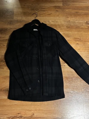 Svart rutig overshirt från Jack & Jones - Säljer en svart rutig overshirt från Jack & Jones i storlek S. Skjortjackan har knappar framtill, två bröstfickor med lock och lång ärm. Perfekt lager på lager-plagg med stilren look och diskret rutmönster. Har en exakt likadan fast svart, båda för 450 eller en för 350.