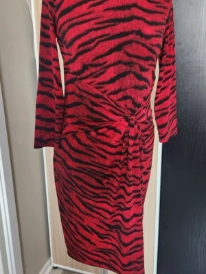 Röd zebramönstrad klänning Michael Kors - Snygg röd klänning från Michael Michael Kors med svart zebramönster. Modellen är knälång med lång ärm och har en cool knytning i midjan. Materialet är mjukt och stretchigt, perfekt för dig som gillar att sticka ut med djurmönster.