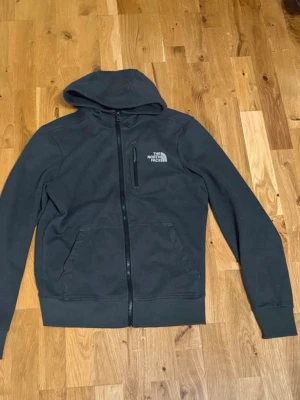 Svart hoodie från The North Face - Svart hoodie från The North Face med huva och hel dragkedja framtill. Tröjan har två stora fickor och en mindre bröstficka med dragkedja. Materialet är mjuk fleece som håller dig varm under kyliga dagar. Perfekt för dig som gillar en enkel och stilren look.
