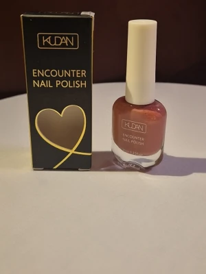 KUDAN Encounter Nail Polish  - Snygg nagellack från KUDAN i färgen brunrosa, levereras i en stilren glasflaska med vit kork. Kommer med originalförpackning i svart och guld. Perfekt för dig som vill ha en elegant och modern look på naglarna.
