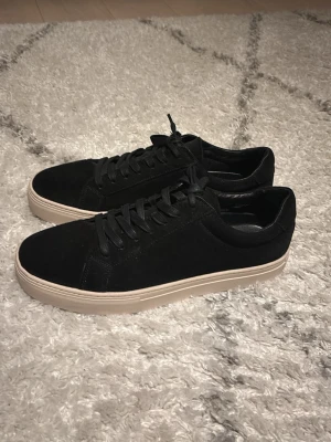 Svarta sneakers från Vagabond - Säljer ett par stilrena svarta sneakers från Vagabond med beige platt sula och snörning. Skorna är tillverkade i mocka och har en klassisk rund tå. Perfekta för dig som gillar enkel och snygg design.           Aldrig använda, helt nya, kartong finns ej kvar!